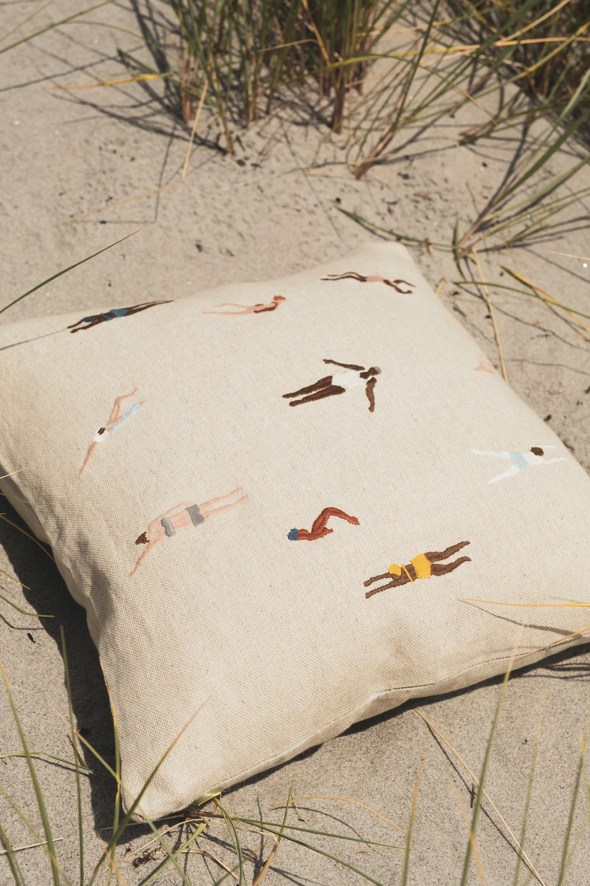 Cojín crema con nadadores bordados en una playa de arena con hierba dispersa.