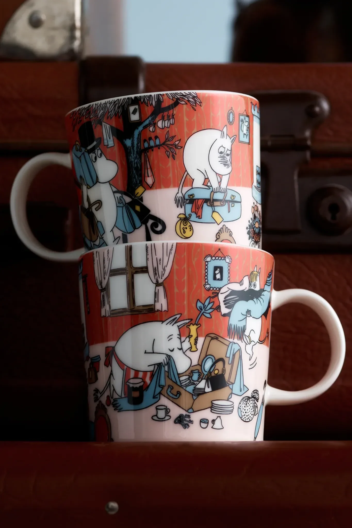Dos tazas Moomin rojas apiladas con ilustraciones de personajes empacando maletas y colgando objetos de un árbol.