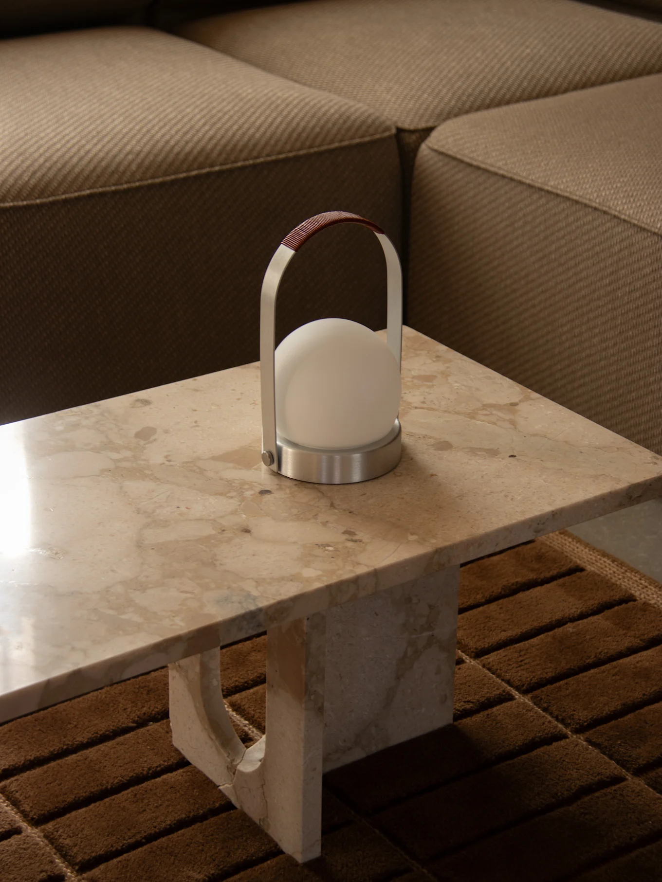 Lámpara portátil moderna con luz blanca esférica, marco plateado y asa de cuero marrón sobre una mesa de mármol junto a un sofá.