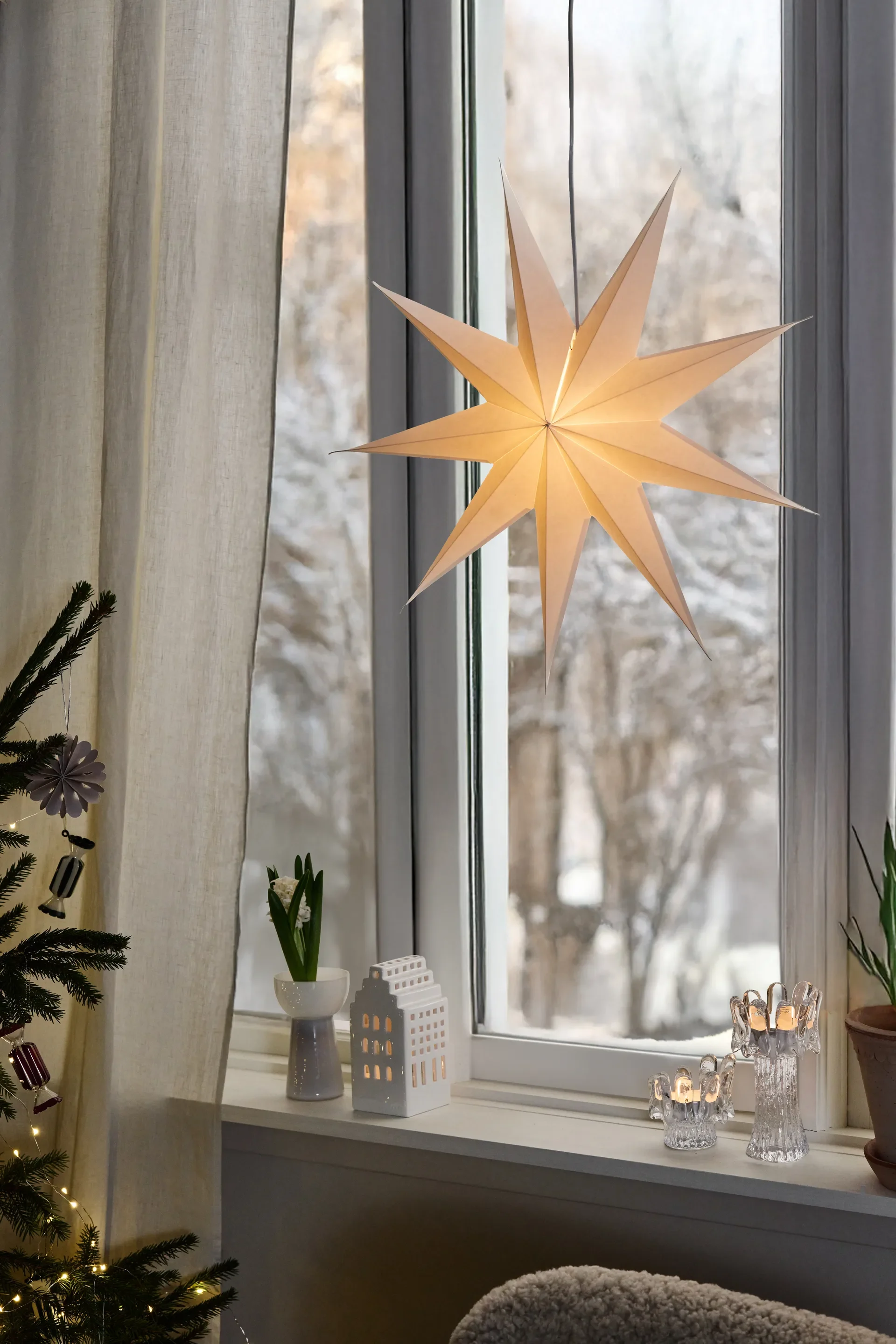 Ventana invernal con estrella de papel brillante, casa de cerámica, portavelas encendidos y árbol de Navidad.