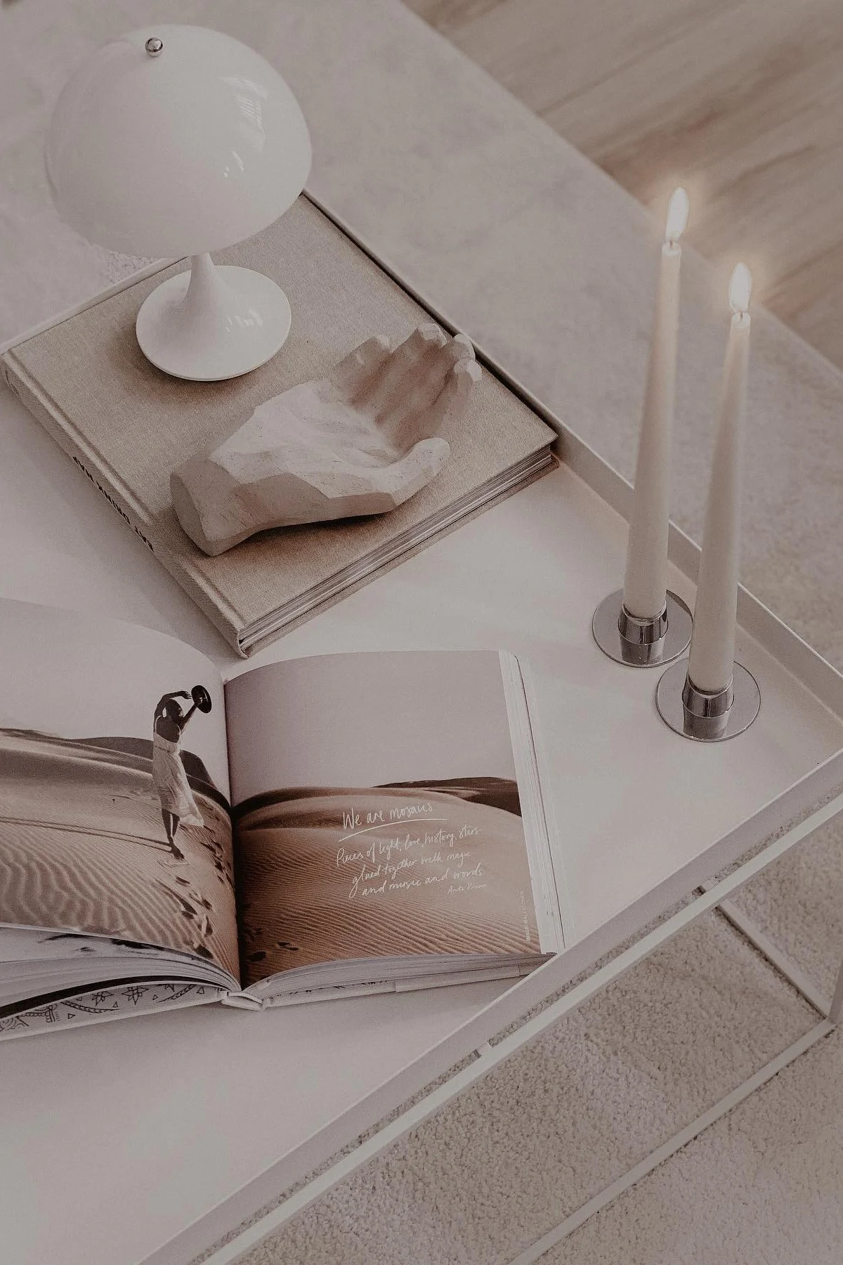 Una mesa de centro amplia, decorada con libros, una mano decorativa, dos velas y una lámpara de mesa portátil Panthella de Louis Poulsen.