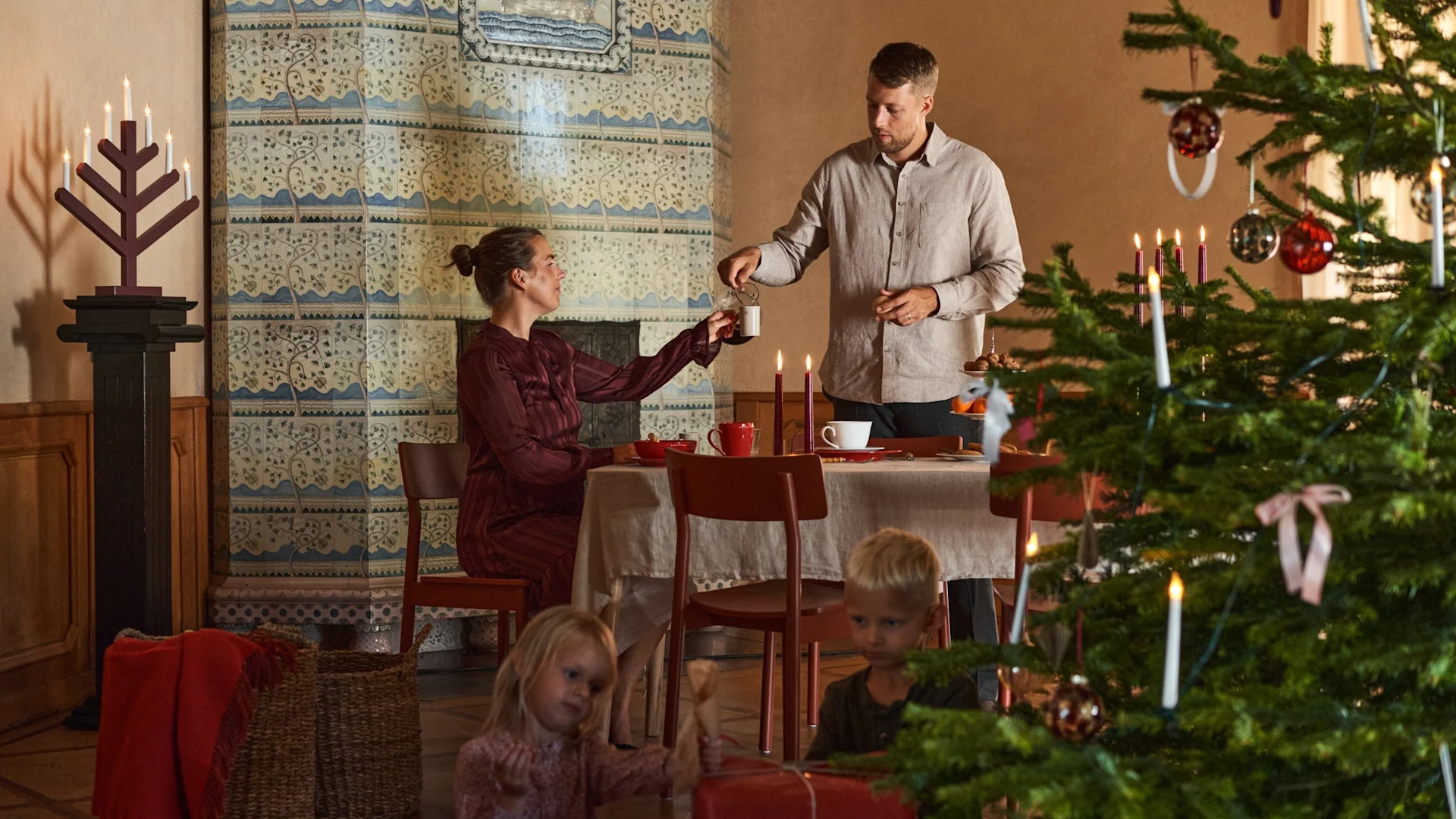 Una pareja está sentada a una mesa decorada para Navidad, junto a un árbol de Navidad con niños que están abriendo los regalos.
