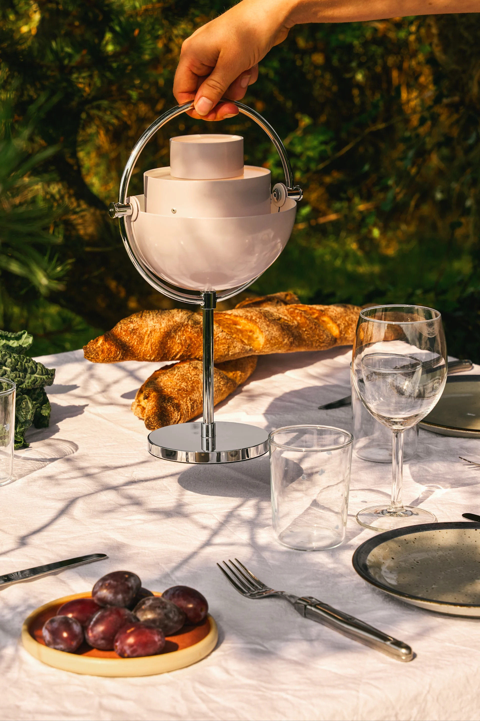 Mano sostiene plato de servir esférico, escalonado y rosa claro con asa de metal en una mesa de exterior.