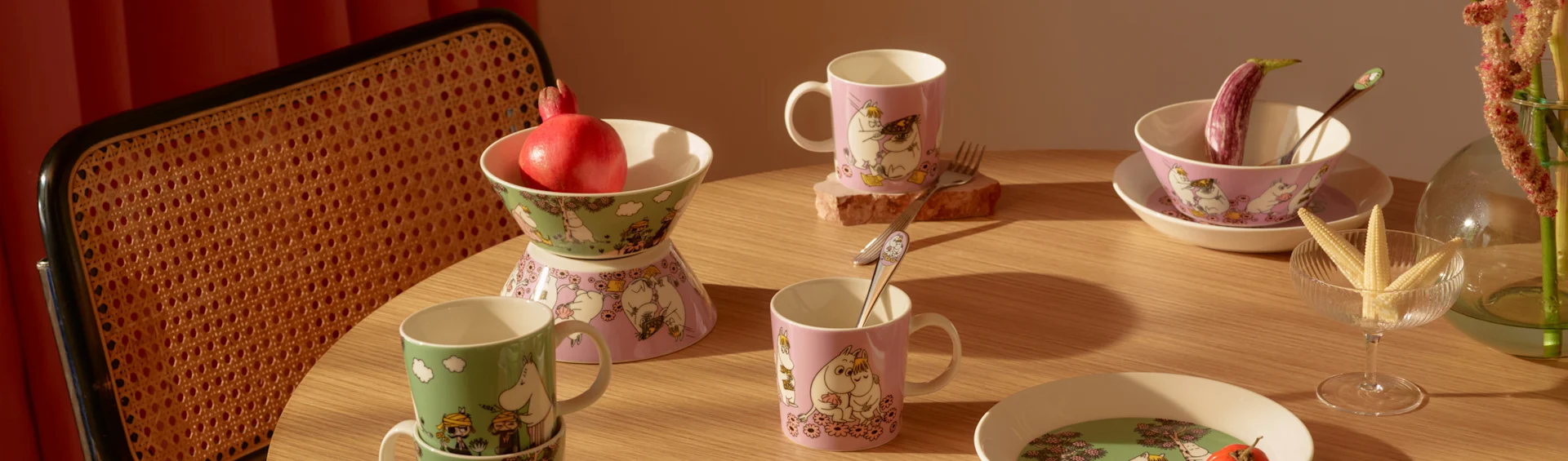Mesa de madera con tazas, cuencos y platos de Moomin verdes y rosas, granada, berenjena y maíz baby.