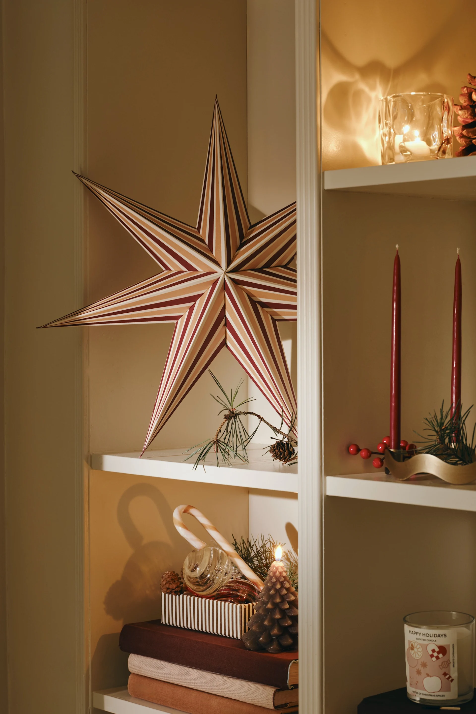 Una estantería blanca decorada con adornos navideños: una gran estrella a rayas, velas cónicas rojas, una piña, una vela con forma de piña encendida, un bastón de caramelo y una vela perfumada con el mensaje «Happy Holidays».