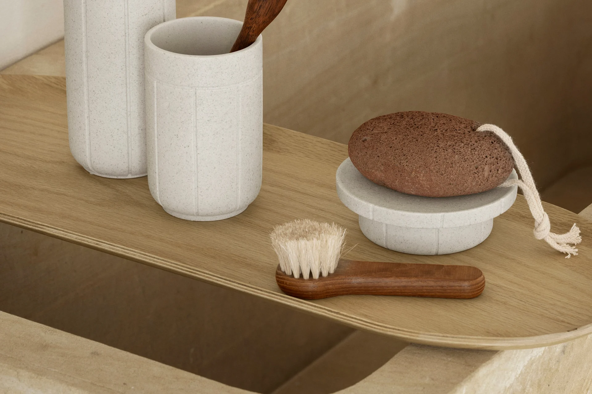 Una bandeja de madera clara con accesorios de baño: dos tazas grises moteadas, una cuchara de madera, una piedra pómez marrón en un soporte y un cepillo de madera.