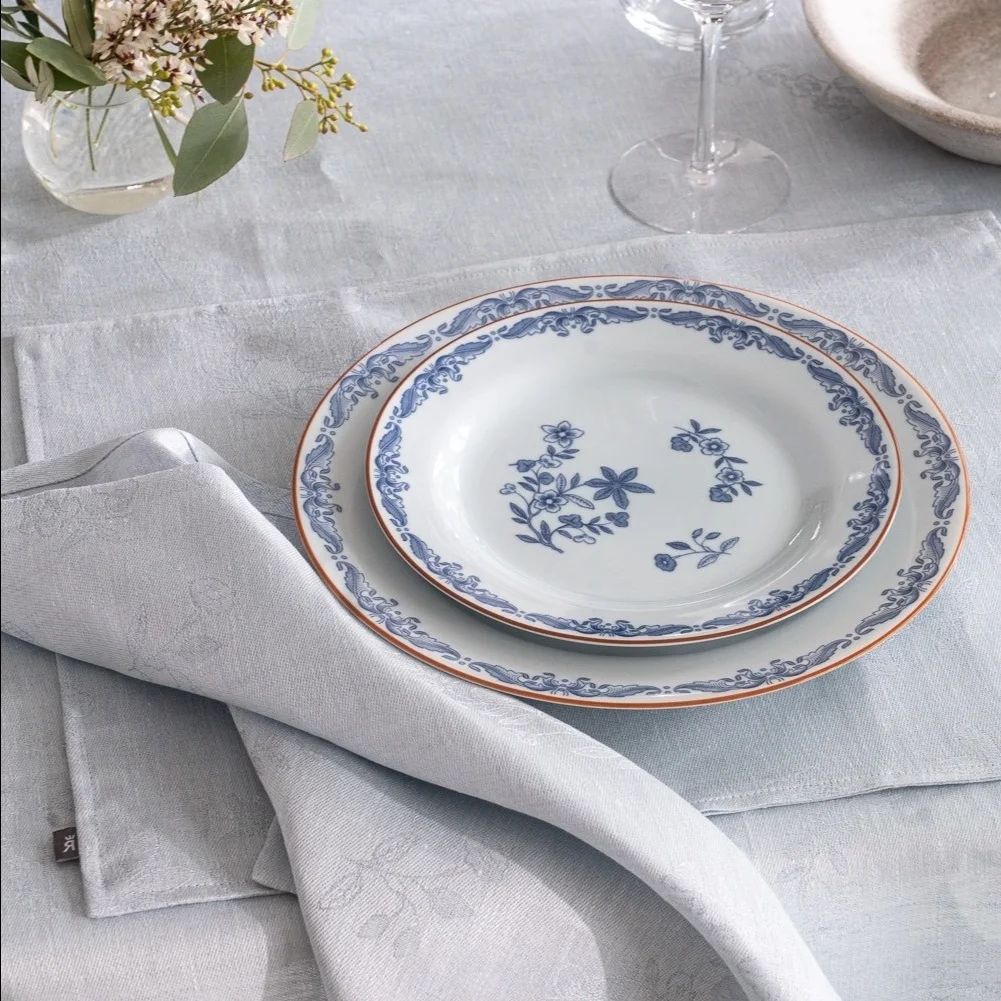 Platos blancos apilados con estampados de flores y ornamentos azules, bajo mantel individual azul claro y servilleta doblada sobre la mesa.