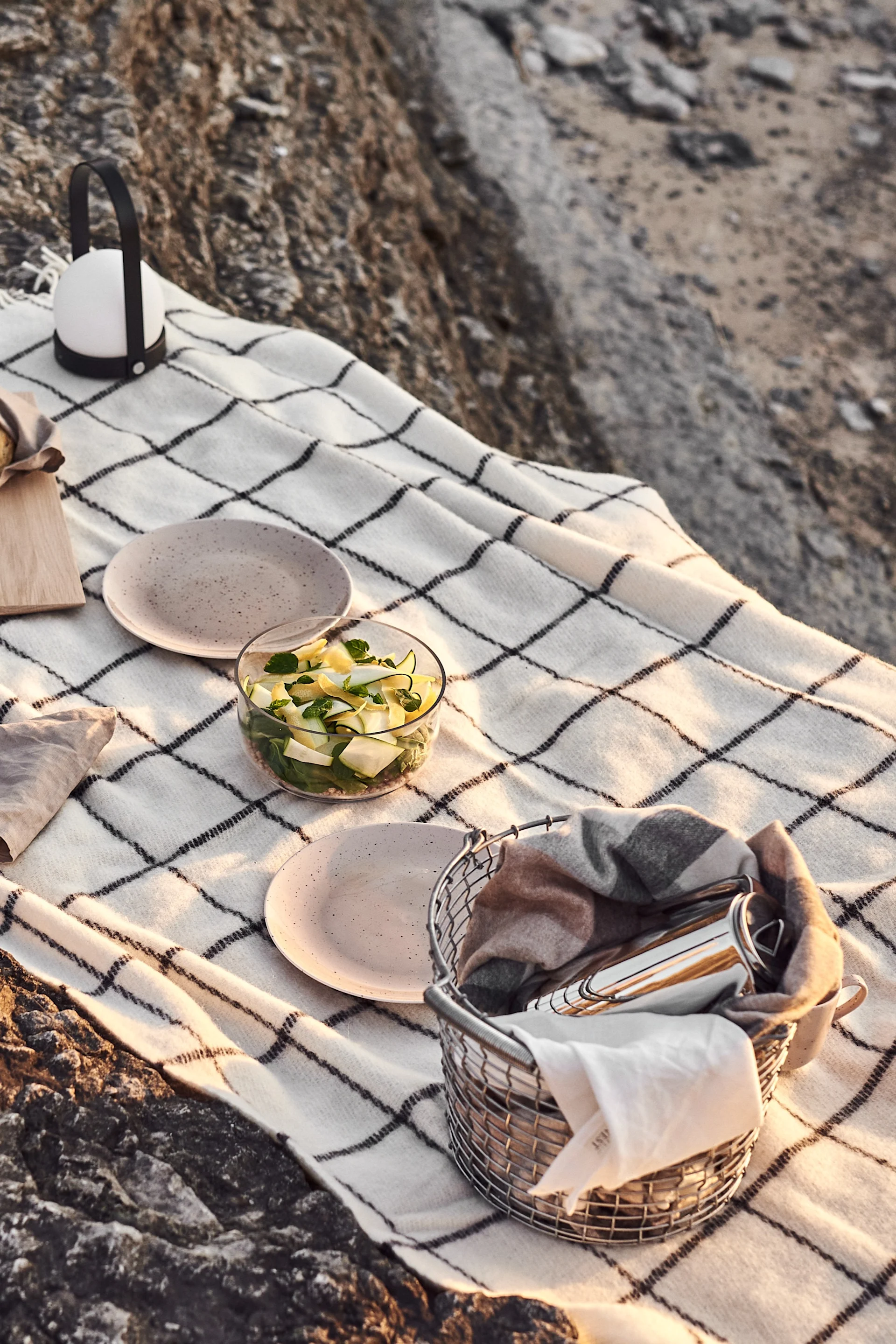 Manta de picnic sobre rocas con ensalada, platos, una linterna y una cesta de utensilios.