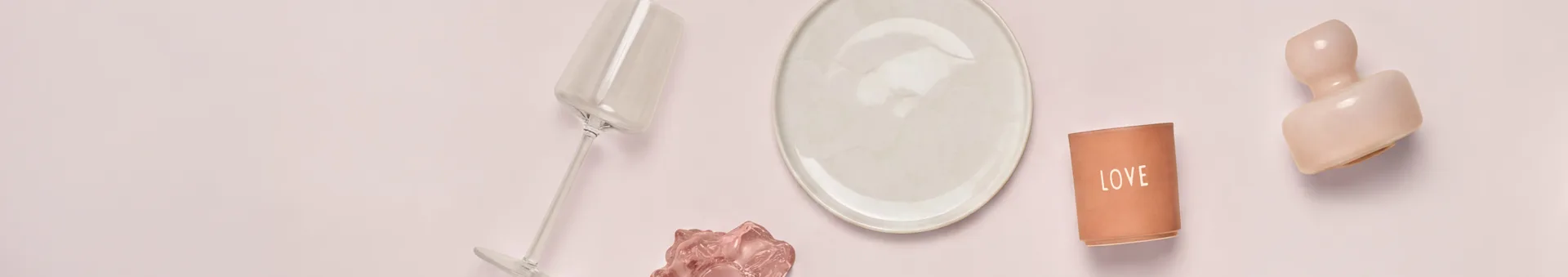 Vista superior de varios objetos decorativos y de mesa sobre una superficie rosa claro. De izquierda a derecha: una copa transparente inclinada, un plato crema, un portavelas de terracota con "LOVE" y una escultura abstracta rosa. Un objeto ondulado rosa más pequeño está debajo de la copa.
