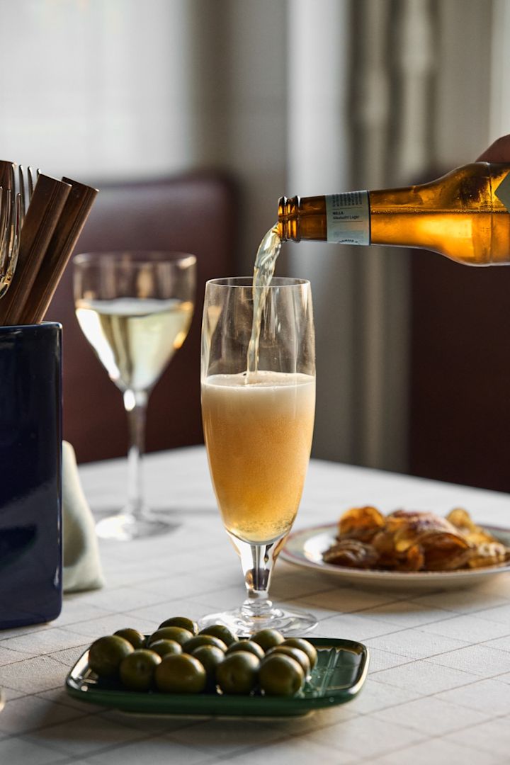 Aprende a elegir el vaso de cerveza adecuado. Aquí puedes ver una cerveza servida en el vaso de cerveza Chateau de Kosta Boda con un plato de aceitunas junto al vaso.