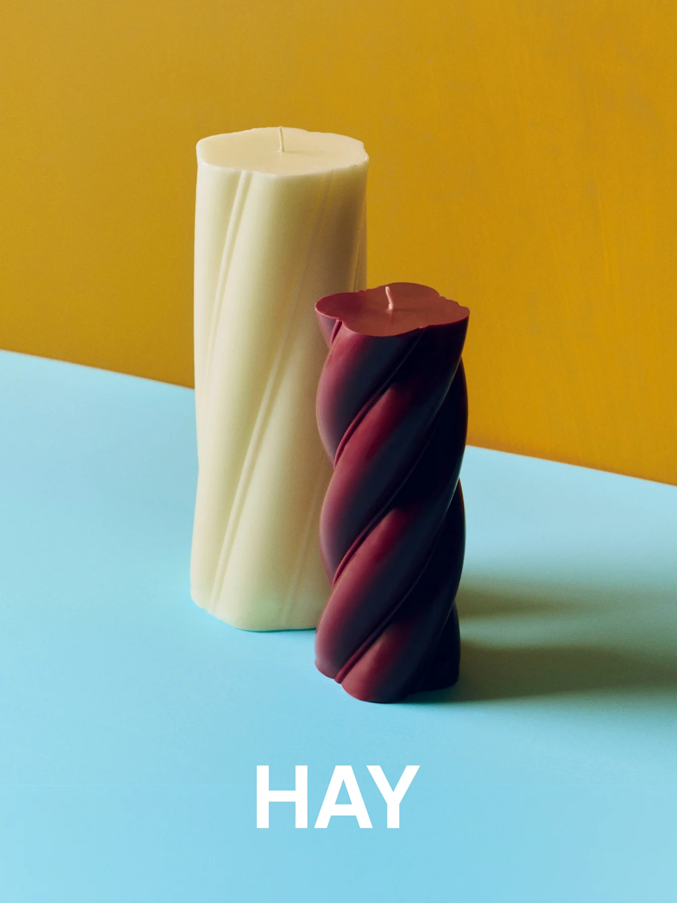 Velas trenzadas de color crema y burdeos sobre fondo amarillo y azul con el texto 'HAY'.