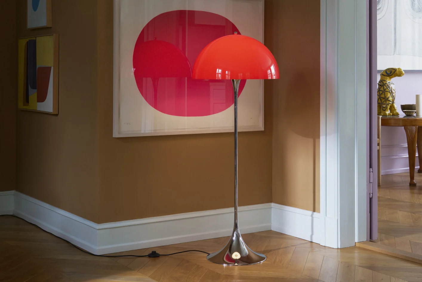 Una lámpara de pie roja brillante con cúpula y pie cromado sobre un suelo de madera en una habitación con paredes marrones y arte moderno.
