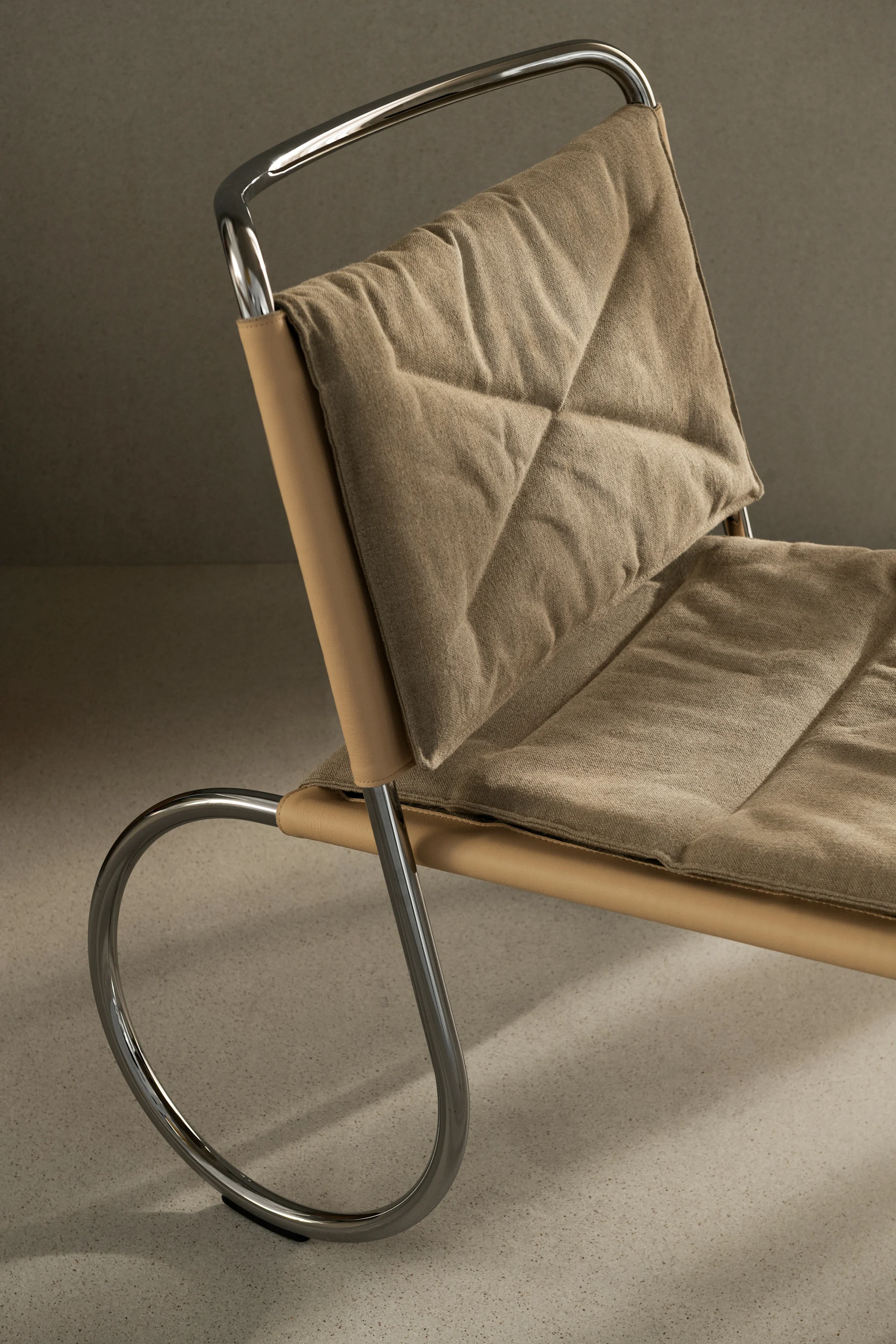 Primer plano de una silla moderna con estructura de cromo pulido, cincha de cuero color canela y cojines de tela beige acolchados.
