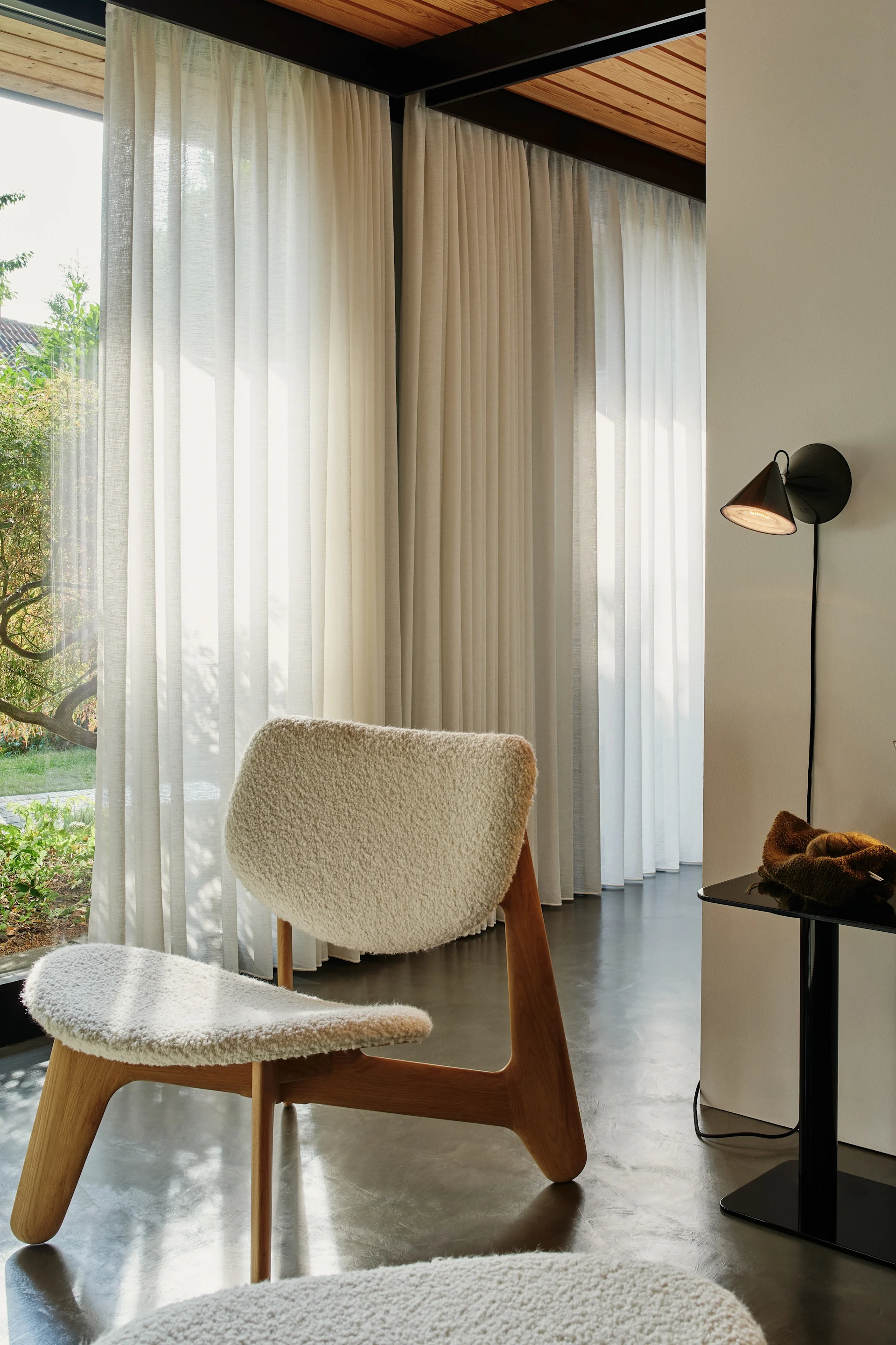 Un sillón de piel de oveja blanca con patas de madera en una habitación luminosa con cortinas transparentes y vistas al jardín. Una lámpara de pared Pose Surface negra de Tom Dixon y una mesa auxiliar. La iluminación de pared es perfecta para decorar paredes. 