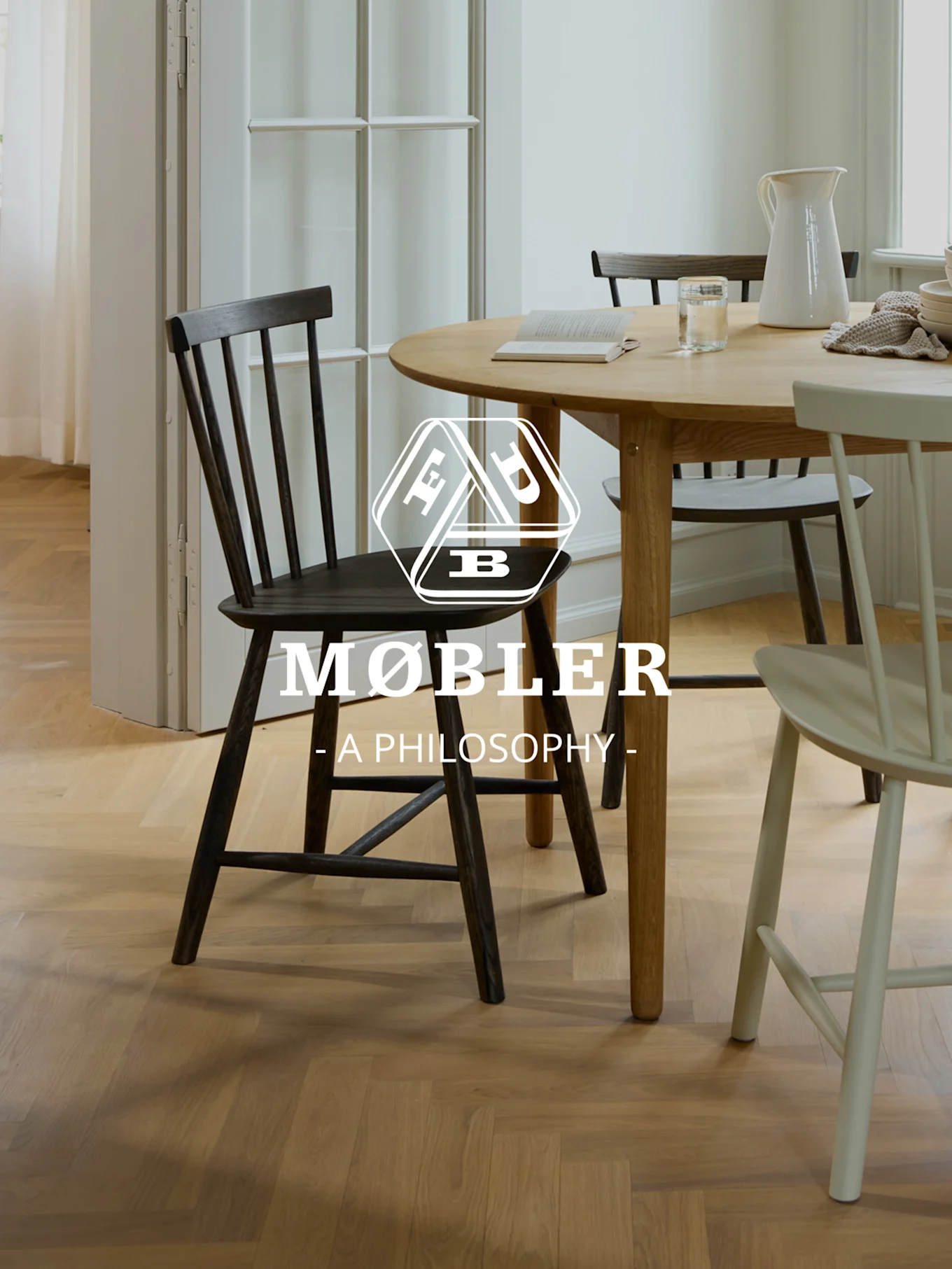 Comedor luminoso con mesa redonda de madera, sillas oscuras y claras, y un logo blanco 'MØBLER - A PHILOSOPHY -'.