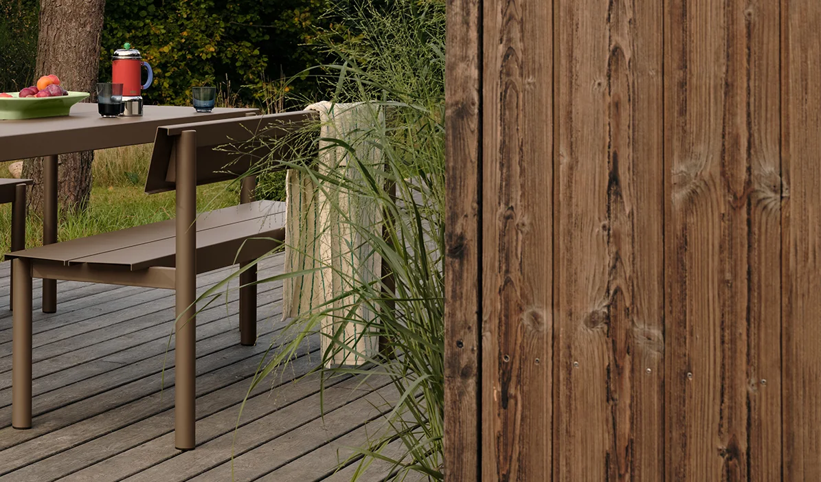 Mesa y banco de metal marrón para exterior en una terraza de madera con frutas y bebidas, junto a hierba alta y una valla de madera.