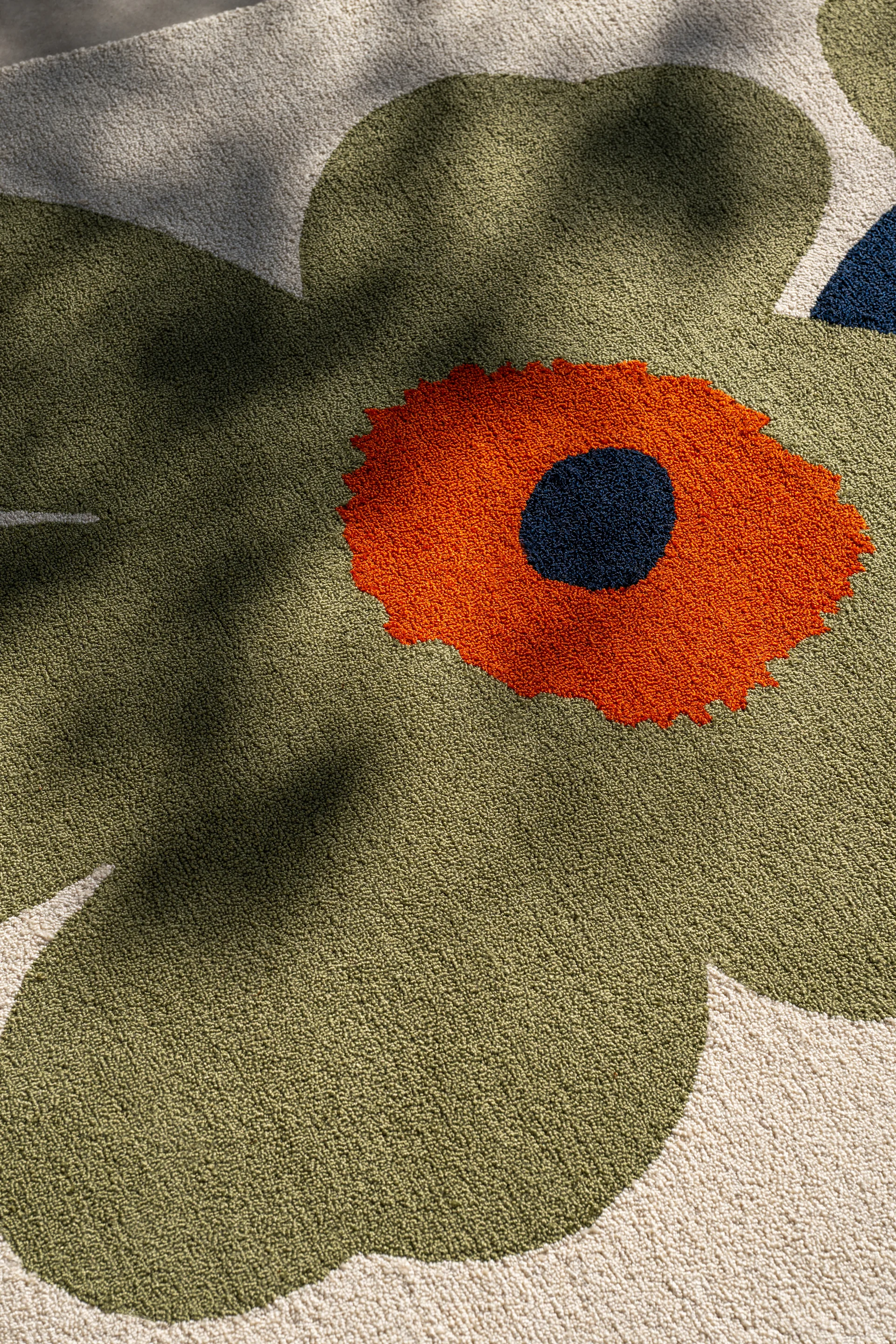 Primer plano de una alfombra con un gran diseño floral estilizado. Pétalos verde oliva, centro naranja y un punto azul oscuro sobre fondo crema.
