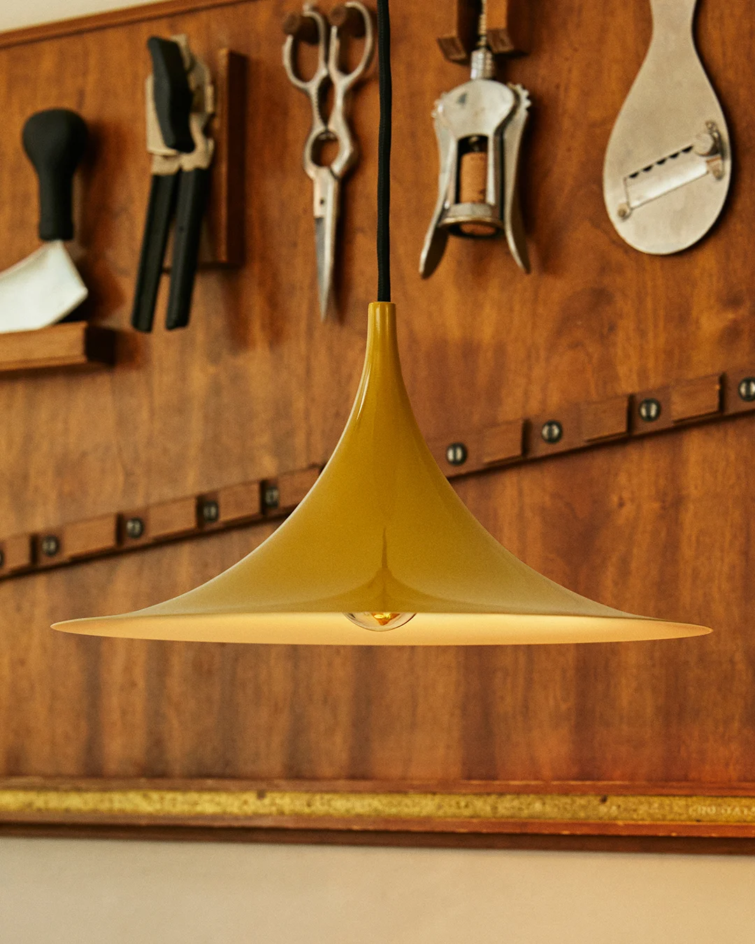 Lámpara colgante dorada en forma de campana con bombilla visible, sobre una pared de madera con herramientas.