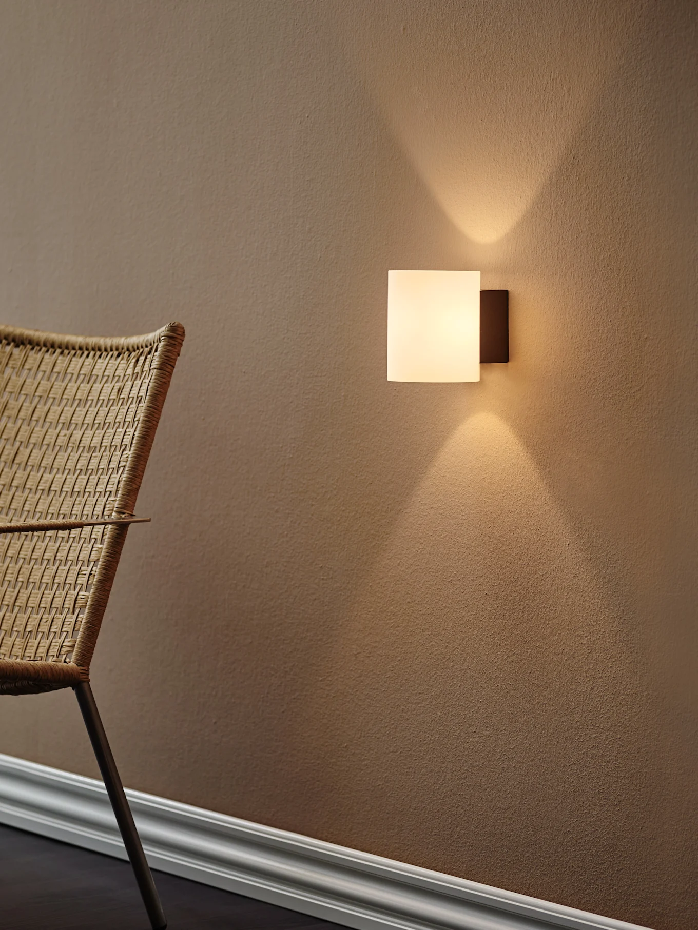 Moderna lámpara de pared cuadrada proyectando luz cálida hacia arriba y abajo sobre una pared marrón texturizada, con una silla tejida a la izquierda.