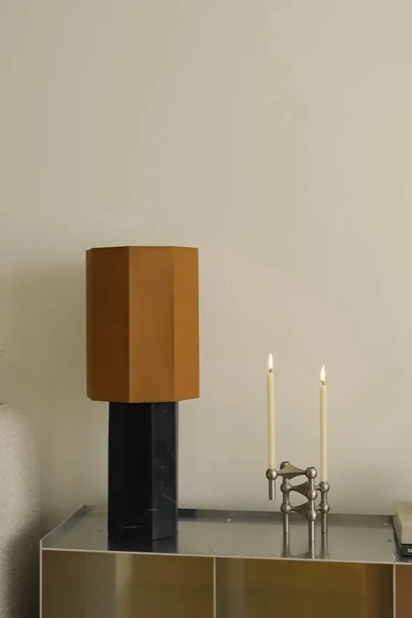 Lámpara de mesa moderna con pantalla hexagonal marrón y base de mármol negro, junto a un candelabro modular plateado con dos velas blancas encendidas.