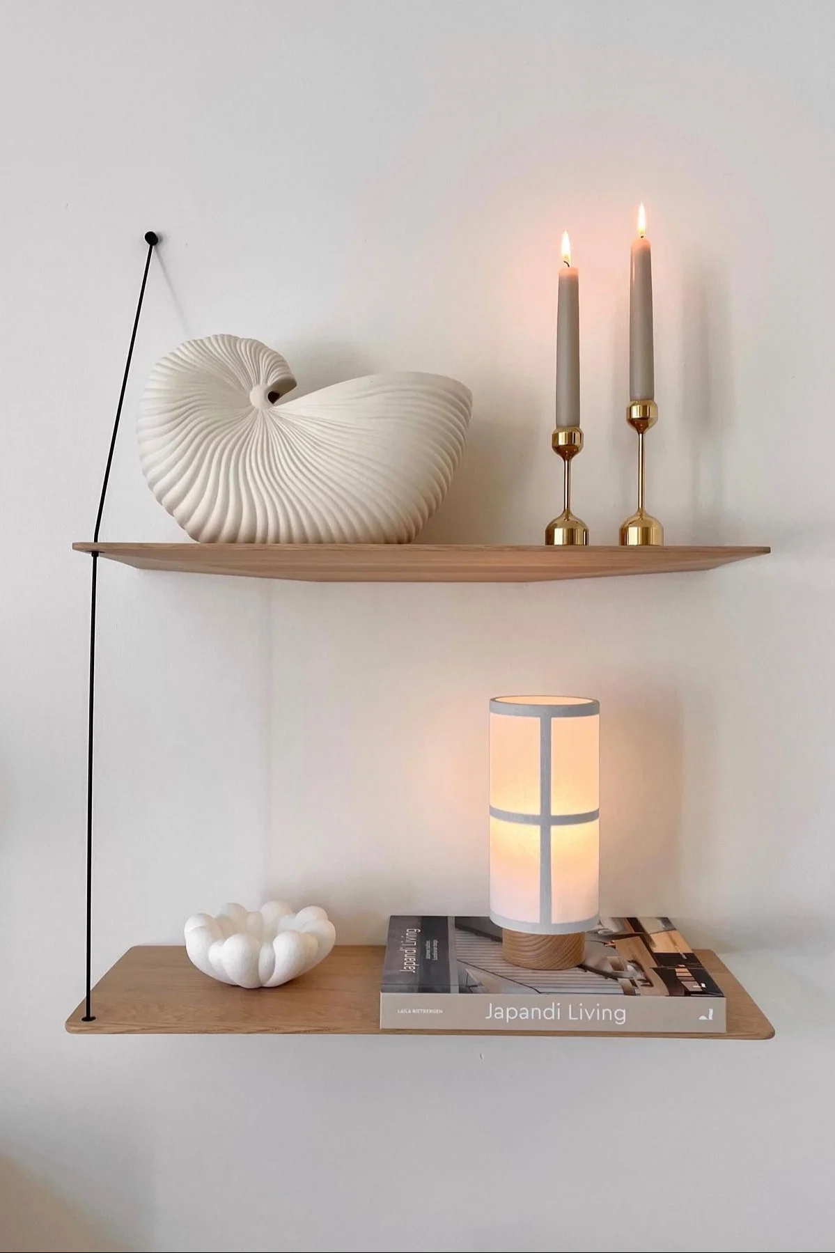 Dos estantes de madera con una concha de nautilus, velas encendidas, un libro Japandi Living, un cuenco con volantes y una lámpara encendida.