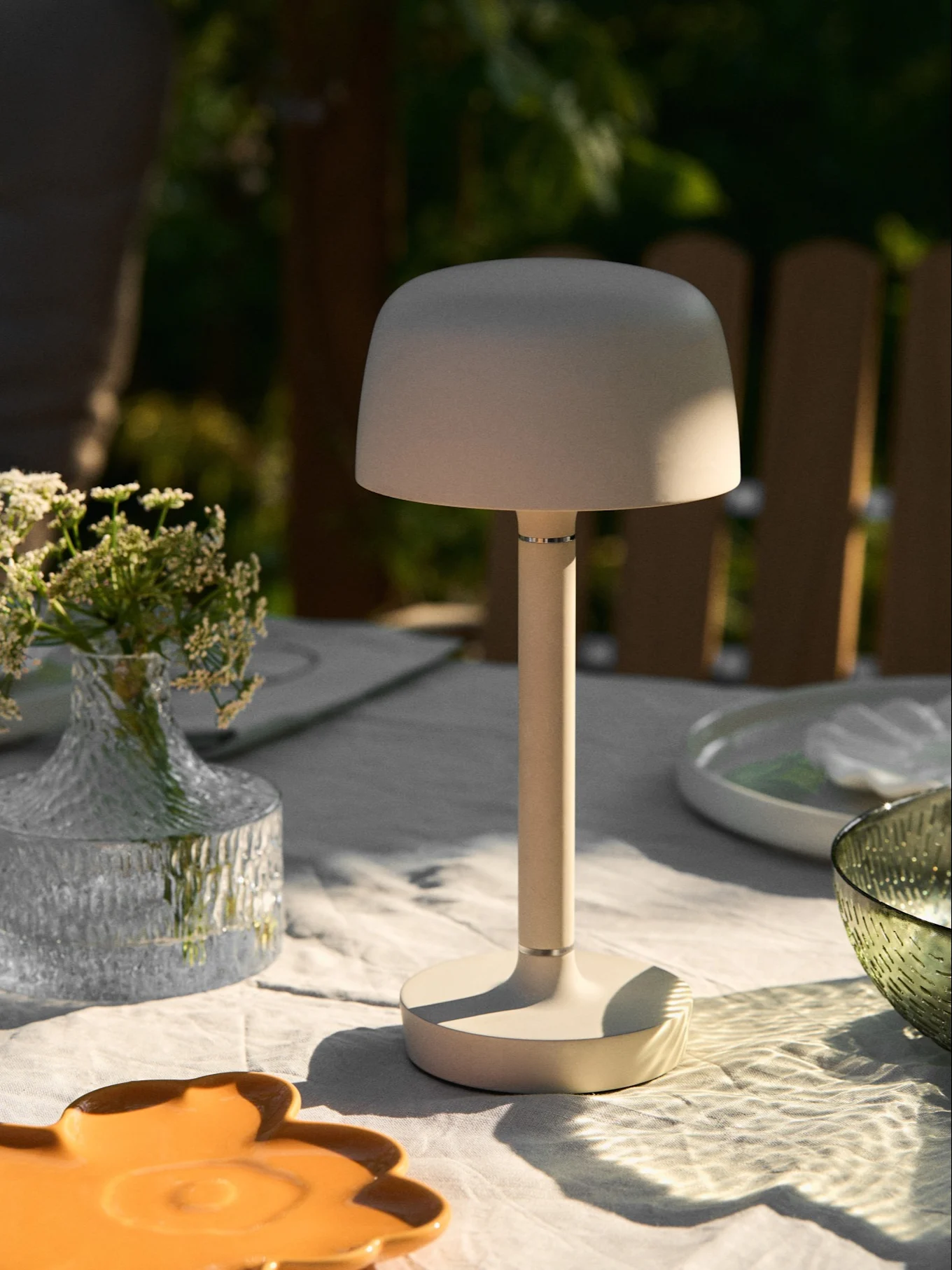 Lámpara de mesa portátil beige en una mesa de exterior con jarrón de flores y luz solar moteada.