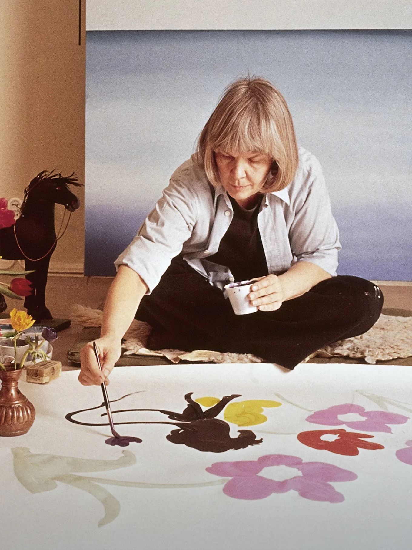 La artista Marimekko Maija Isola sentada en el suelo pintando flores grandes y coloridas sobre una tela blanca.