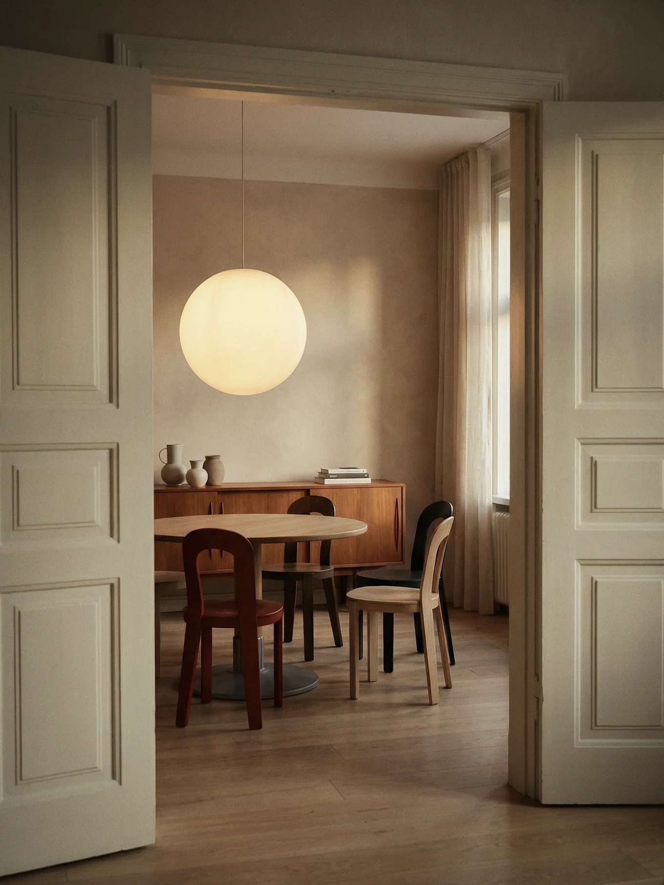 Comedor por puerta abierta, con mesa redonda de madera, sillas variadas, lámpara esférica y aparador de madera.