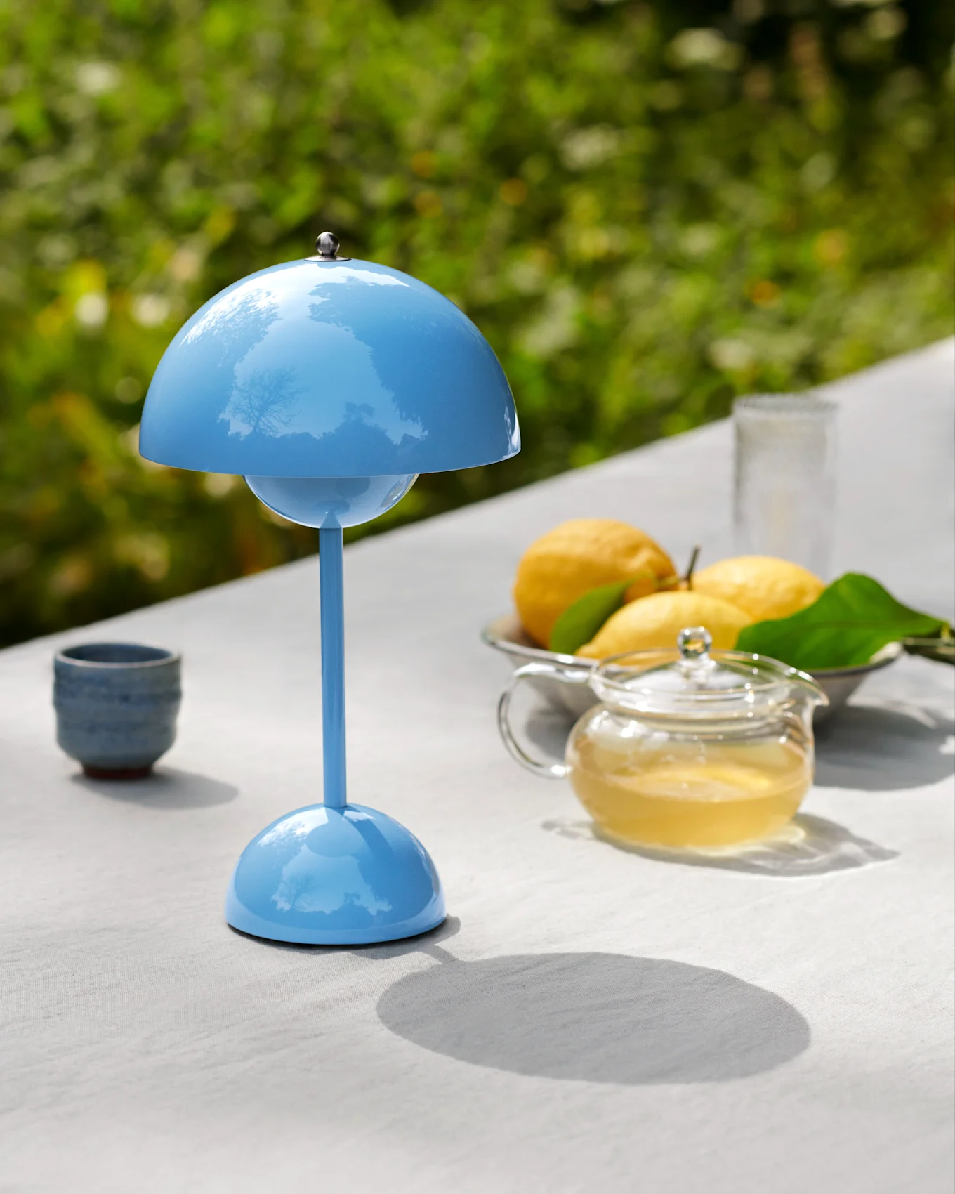 Una lámpara de mesa azul con forma de hongo en una mesa exterior con una tetera, limones y una taza.