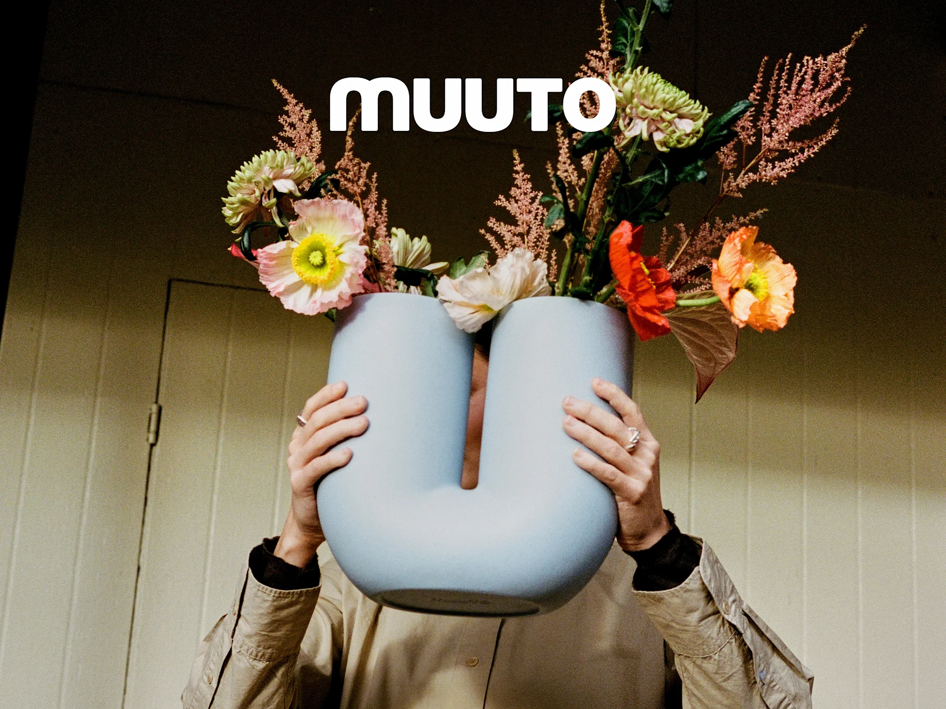 Persona sosteniendo un jarrón azul claro en forma de U con un vibrante ramo de flores rosas, naranjas y verdes. Logotipo de Muuto.
