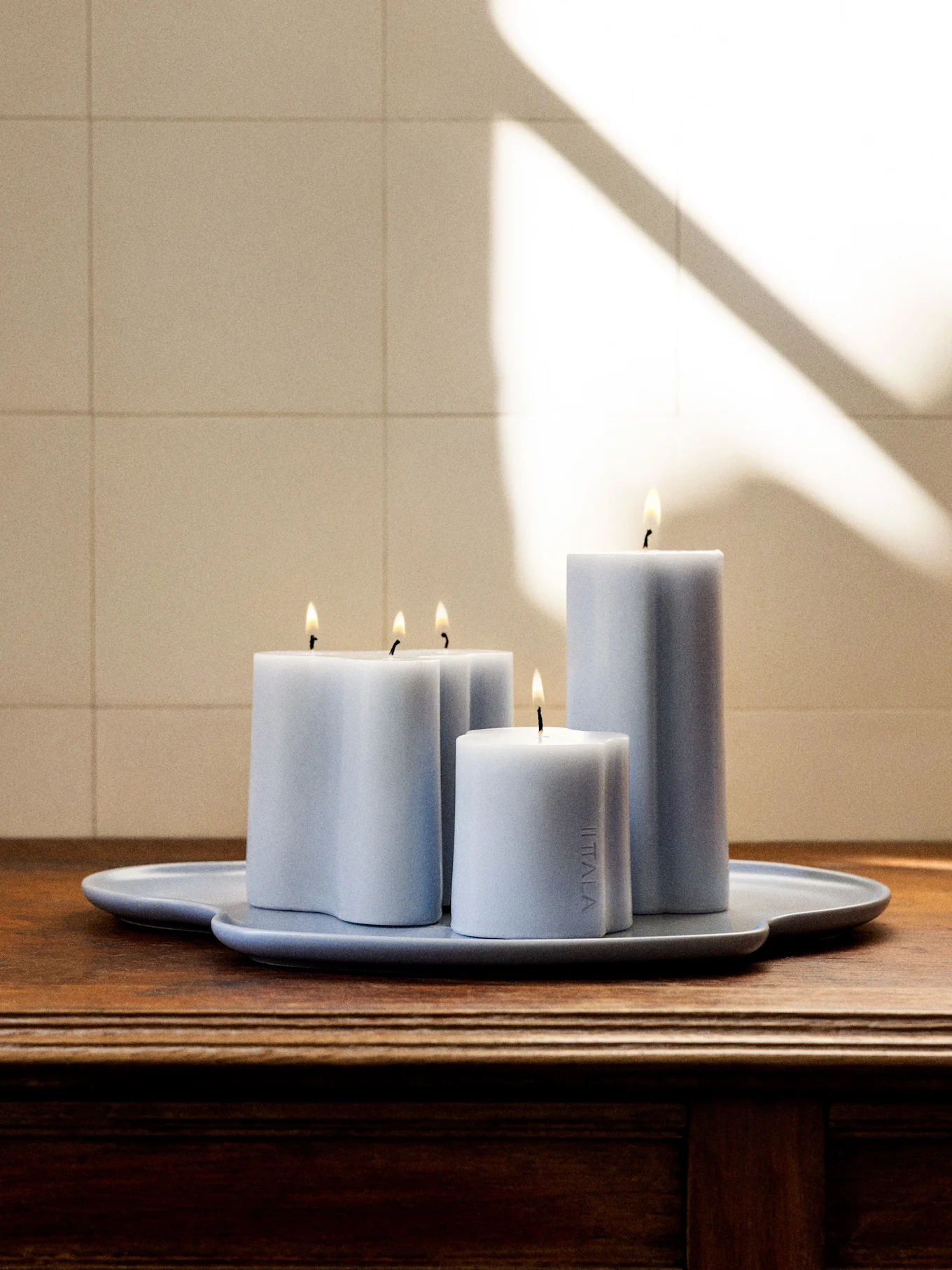 Cuatro velas encendidas de color azul grisáceo claro de varias formas y alturas en una bandeja a juego de forma irregular sobre una superficie de madera oscura.