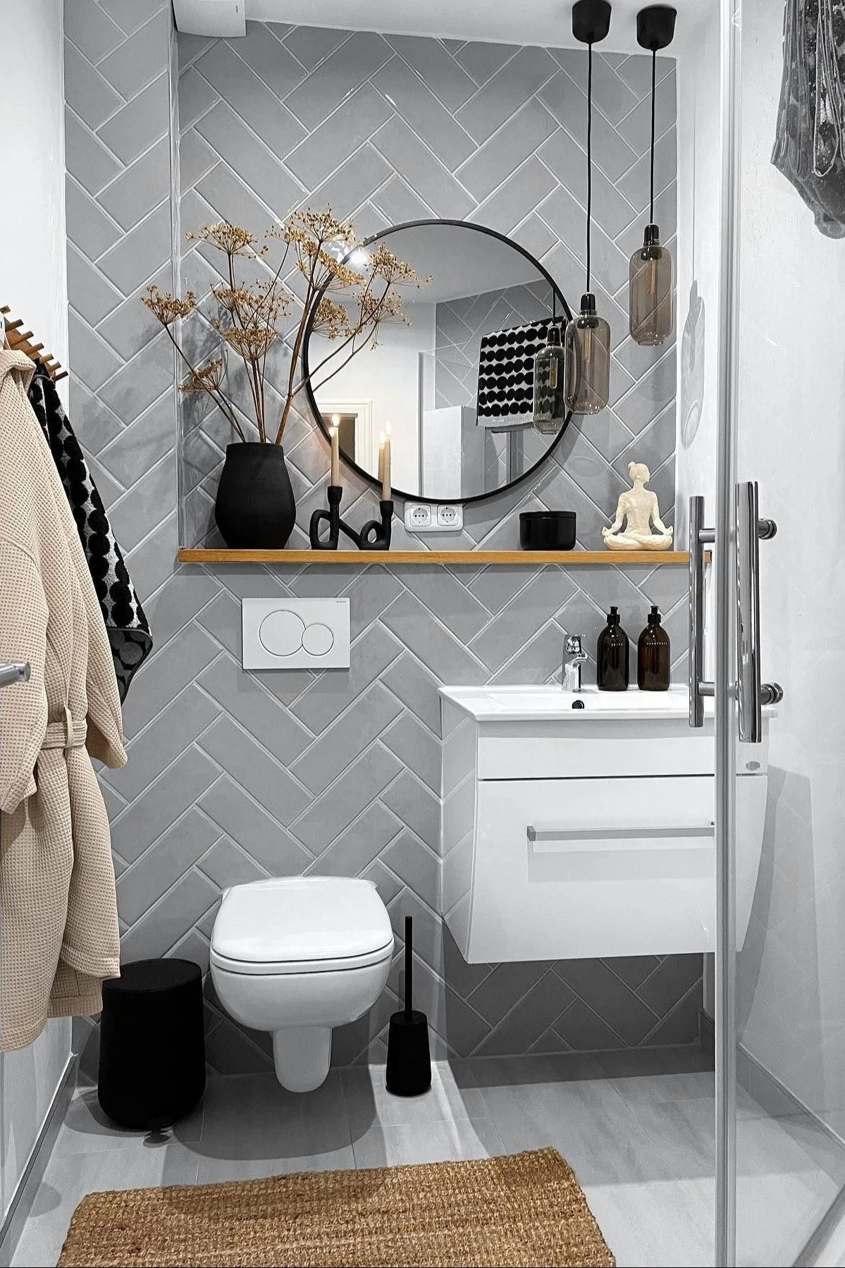 Baño moderno con azulejos de espiga gris claro, inodoro blanco, tocador, espejo redondo y estante de madera con decoración.